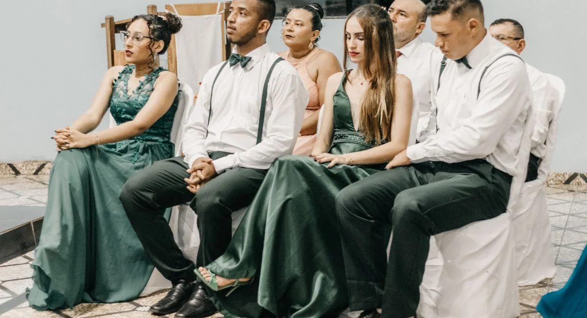A group of elegantly dressed adults sitting attentively indoors at a wedding.