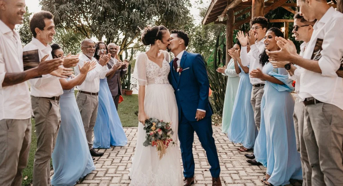 Young bride with flower bouquet kissing black husband between cheerful clapping guests on tiled walkway during festive event