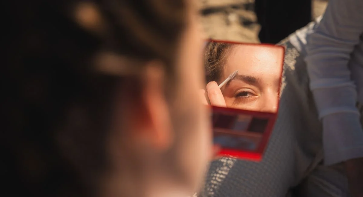 A woman uses a mirror to apply eyebrow makeup outdoors, captured in warm natural light.