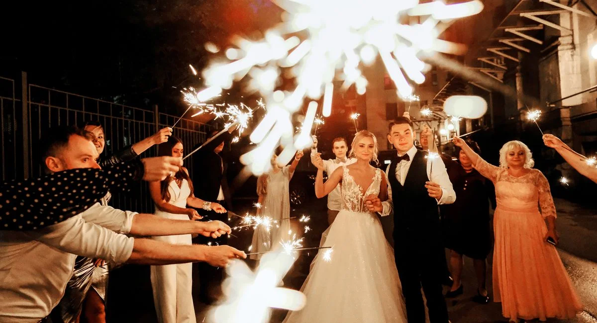 Happy young bride and groom standing on street near unrecognizable guests with burning sparklers during wedding party in even