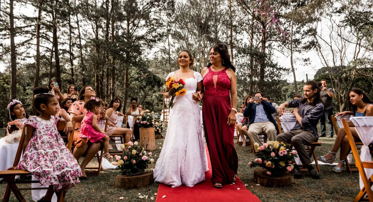 A joyful bride walking with her mother on a red carpet at an outdoor wedding ceremony surrounded by guests.