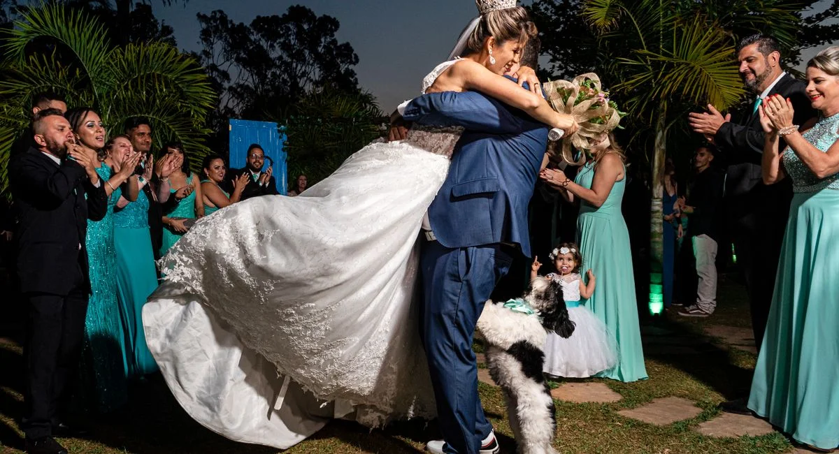 A bride and groom embrace amid a joyful outdoors wedding celebration with happy guests and a dog.