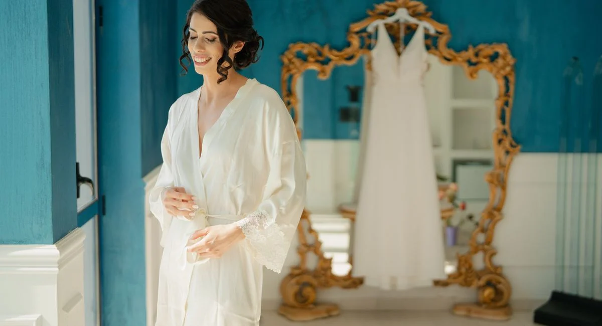 A bride in a robe smiles while preparing for her wedding in a stylish blue room with a gold-framed mirror.