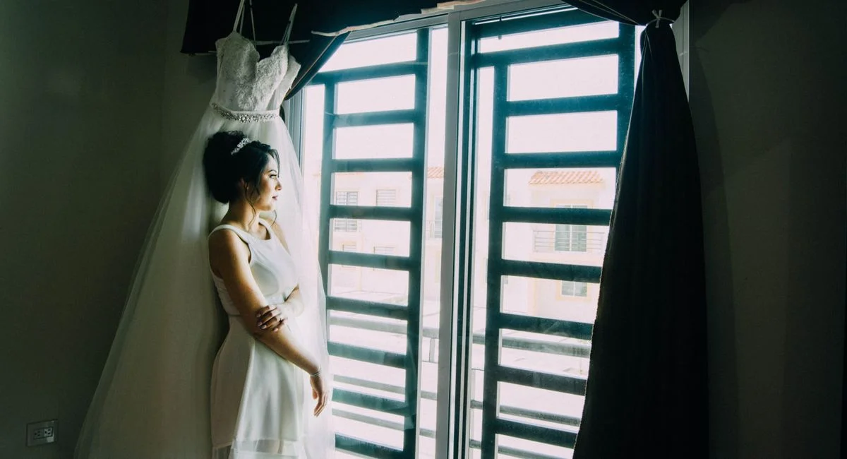 Bride stands by window in white dress, reflecting on her special day. Captures intimate wedding moment.