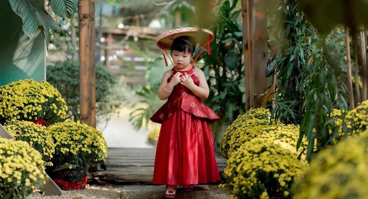 A young girl in a red dress and traditional hat walks through a vibrant flower garden.