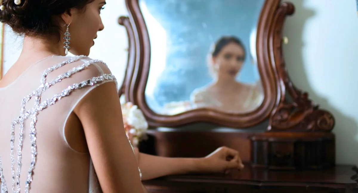 Bride in ornate dress glancing in a vintage mirror, showcasing elegance and anticipation.